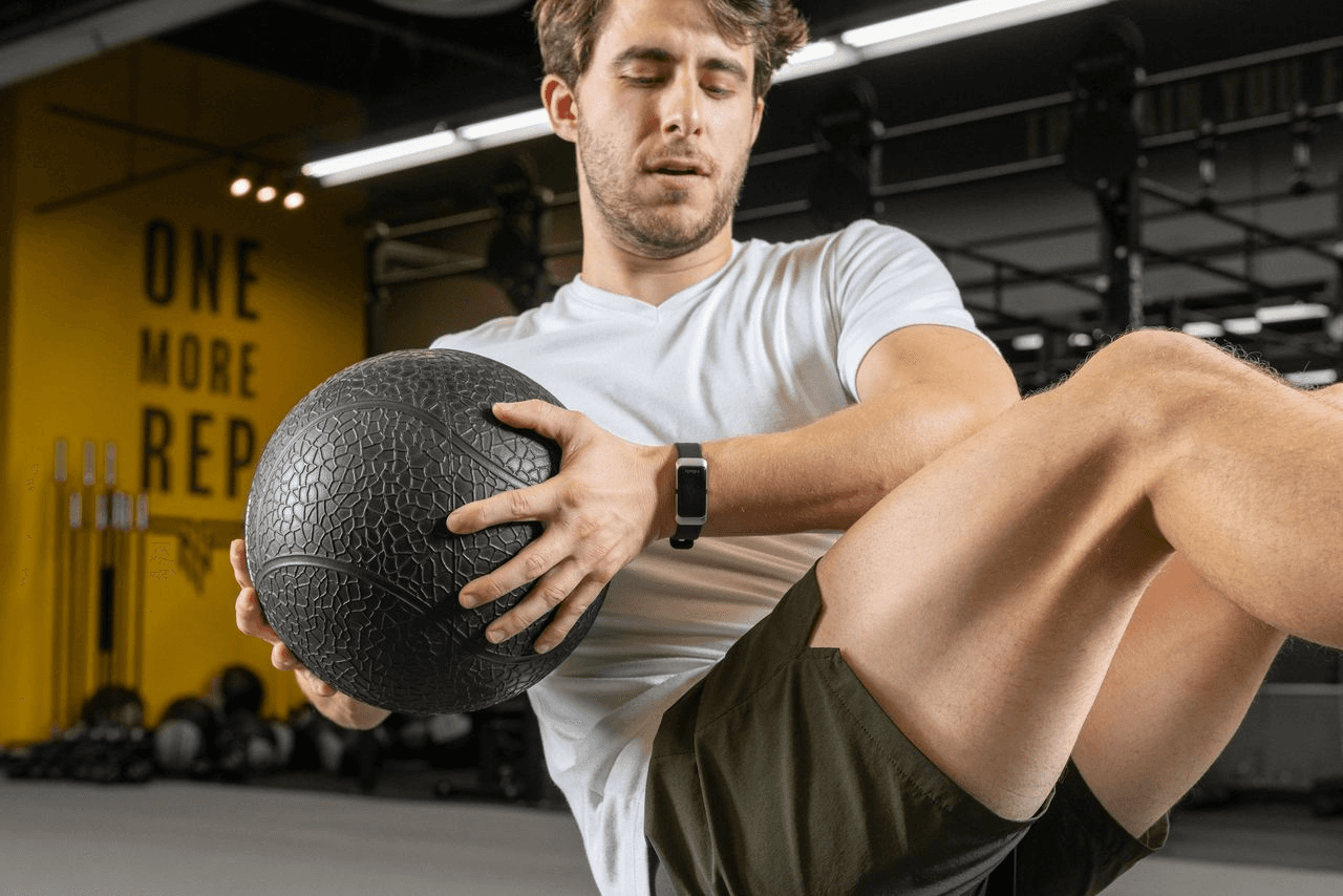A man performing a medicine ball Russian twist exercise in a modern gym with "ONE MORE REP" signage in the background.