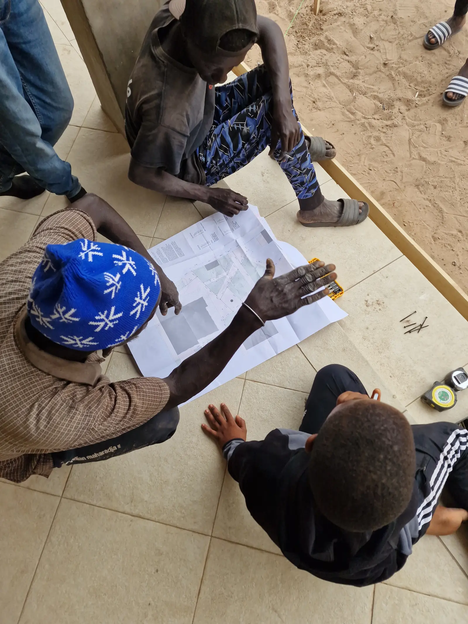 Three individuals seated on the ground, looking at documents and discussing in an informal setting.