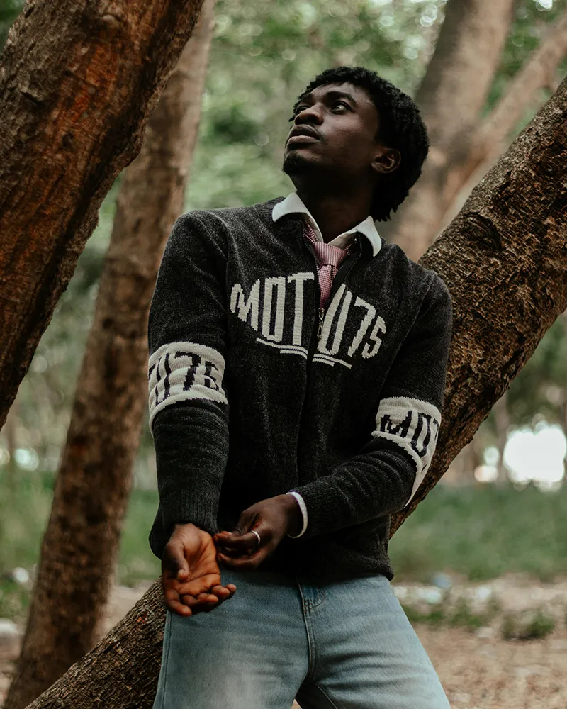 A young man in a graphic sweater looks up while leaning against a tree