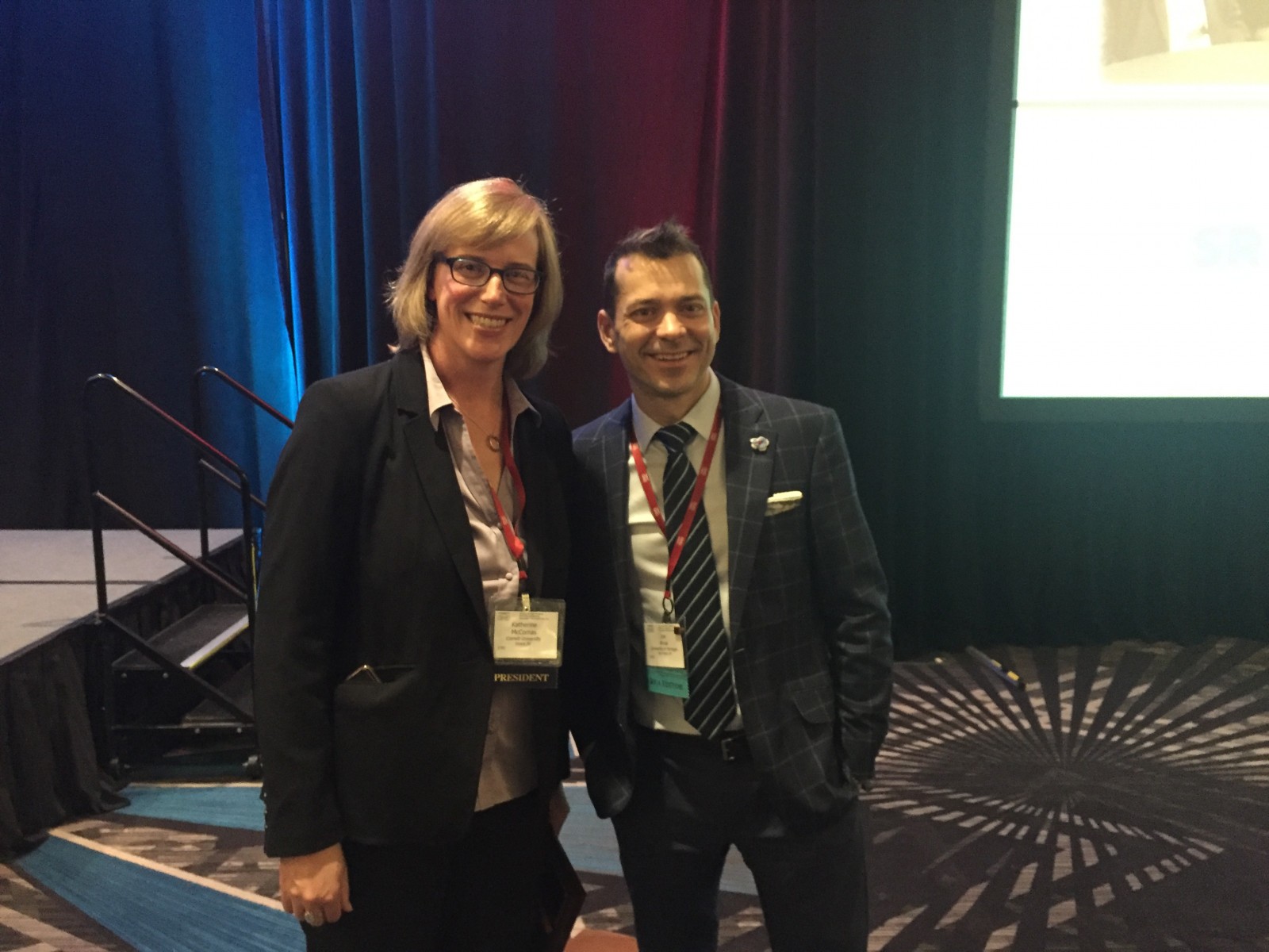 Two individuals smile for the camera at a conference, with colorful lighting in the background.