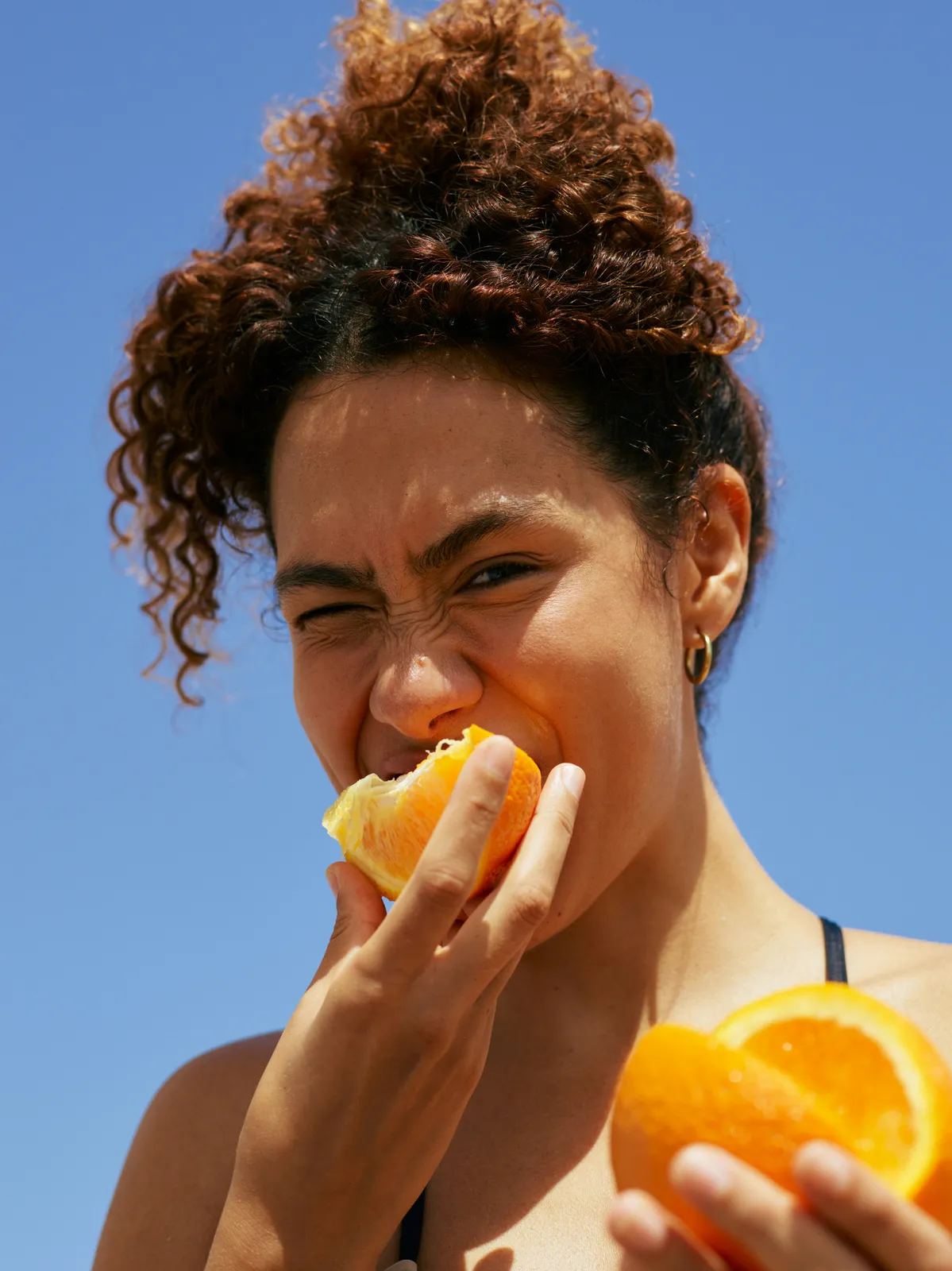 Woman with curly hair biting into an orange slice under a blue sky