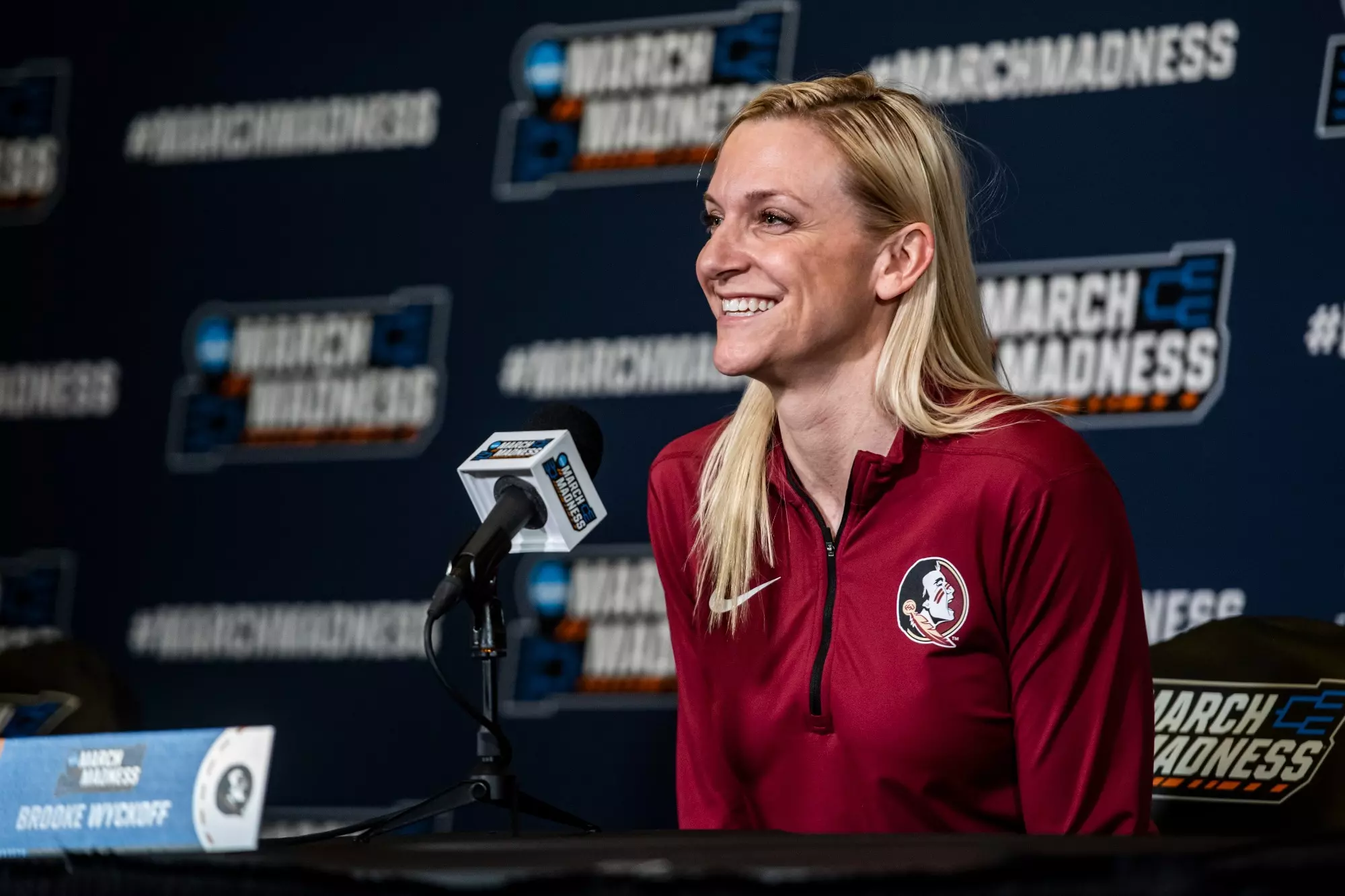 Brooke Wyckoff in a red Florida State Seminoles jacket smiles while speaking at a press conference. She sits in front of a microphone and a backdrop with March Madness logos.