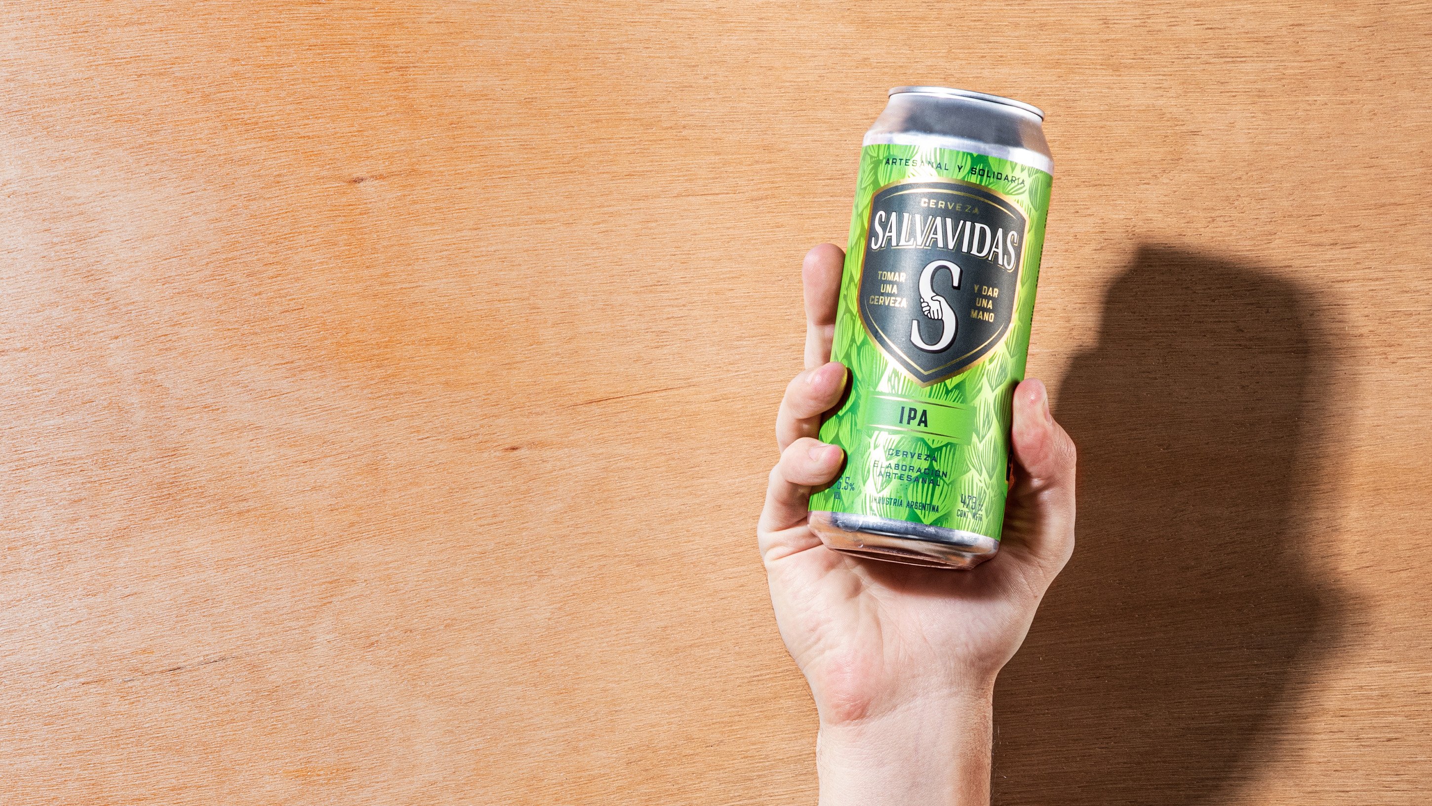 A hand holding a green can of Salvavidas IPA beer against a light wood background. The can's design is vibrant with bold branding and a refreshing tone.