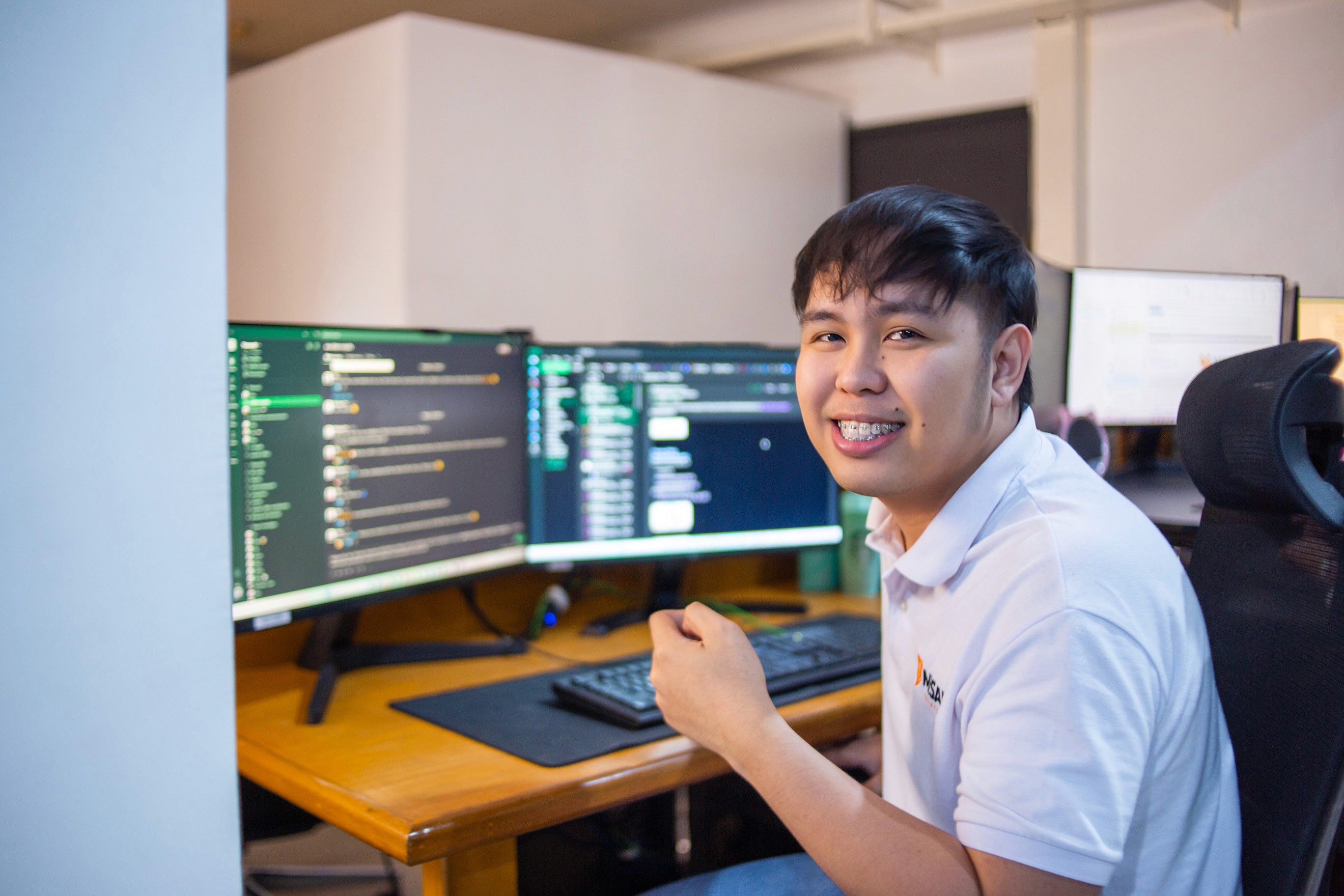 A young man in a white shirt looks up from his computer with a smile, seated at a desk with code on the screen.