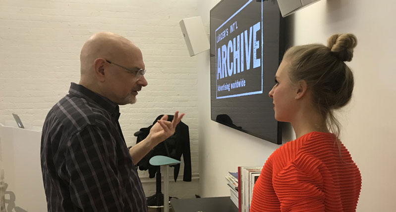 A photo of two people talking in front of a wall mounted tv screen.