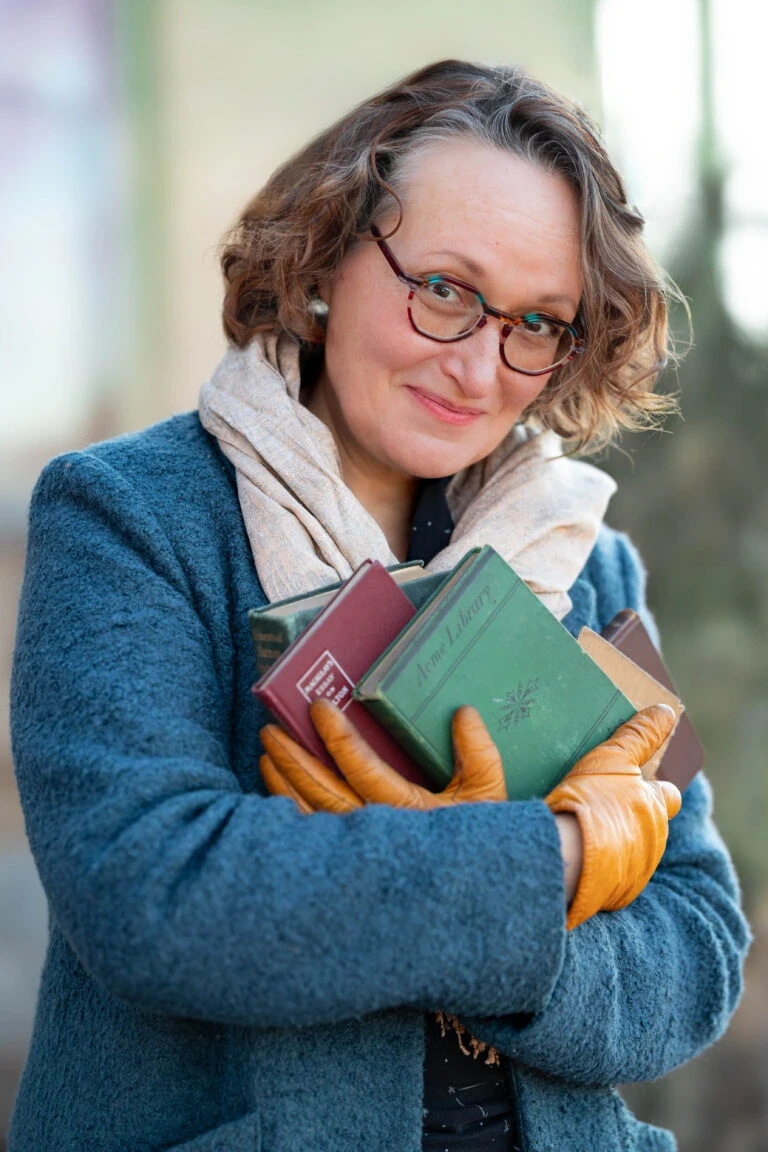 A person in a blue coat and glasses holds several vintage books while standing outdoors, wearing orange leather gloves and a beige scarf, with a blurred background of greenery and a building.