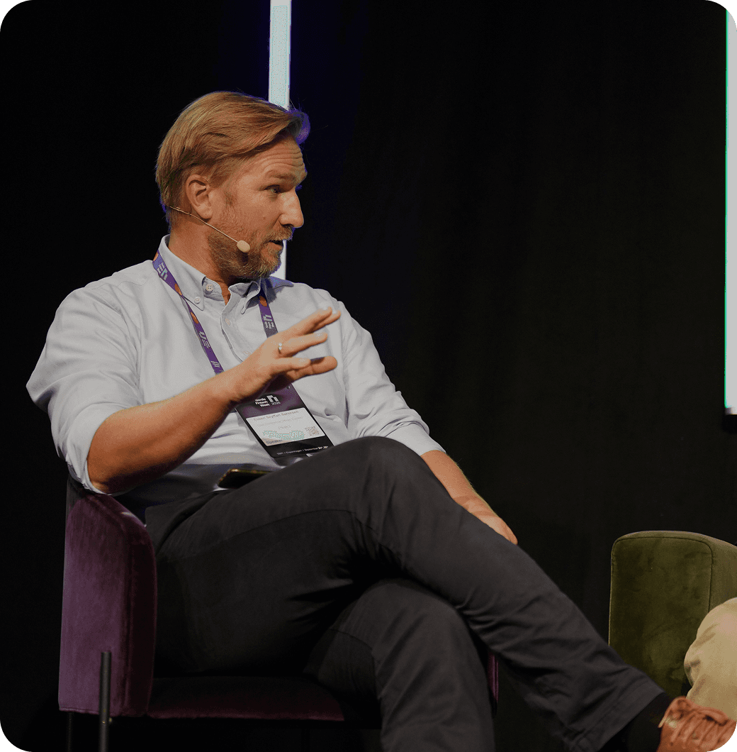 A person seated on a stage, gesturing while speaking during a discussion, with a dark backdrop and colorful seating.