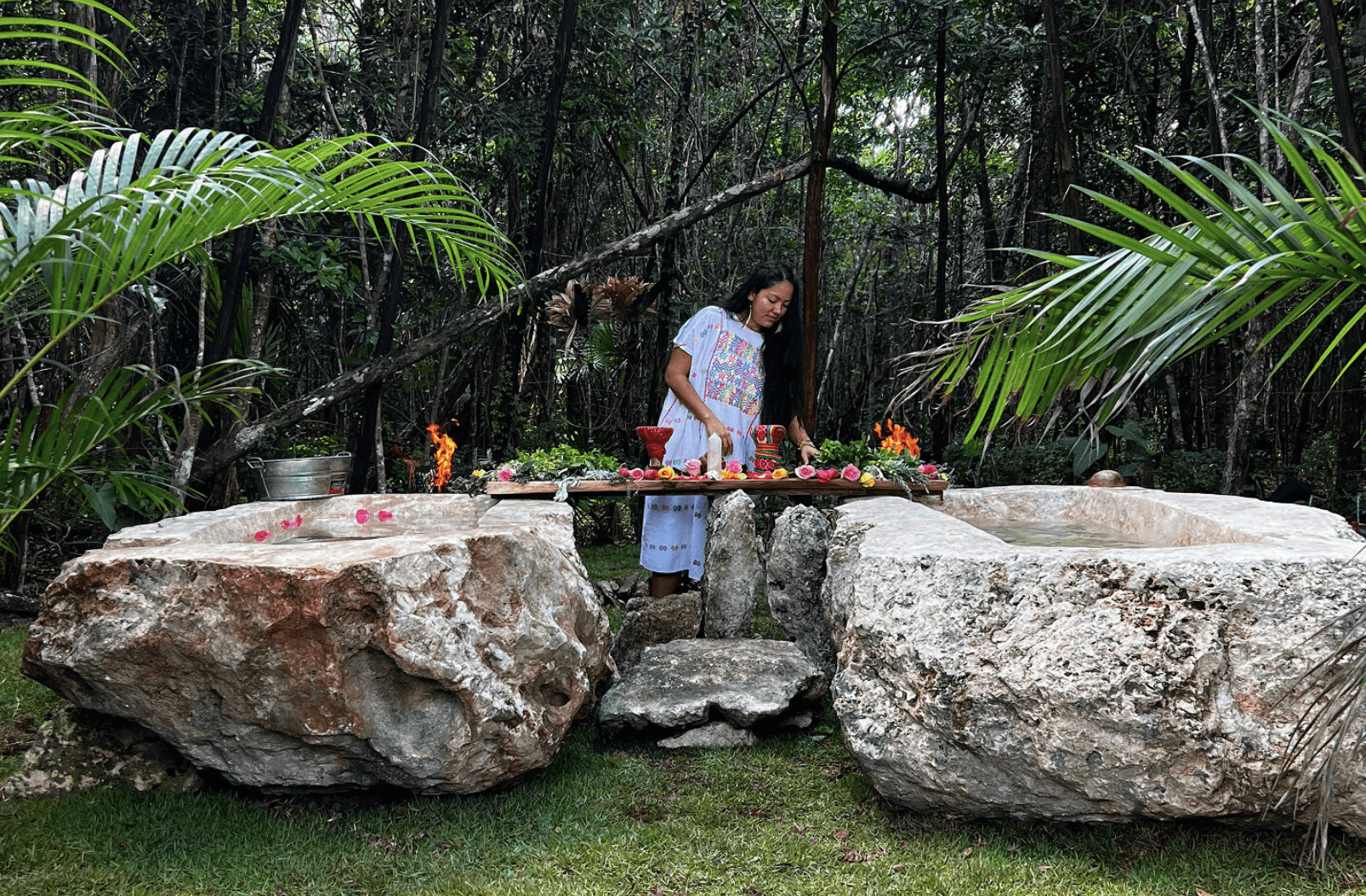 A rustic outdoor market stall displays an array of fresh produce including vibrant tomatoes, leafy greens, and a variety of olives, set against the backdrop of a picturesque rural Italian village.