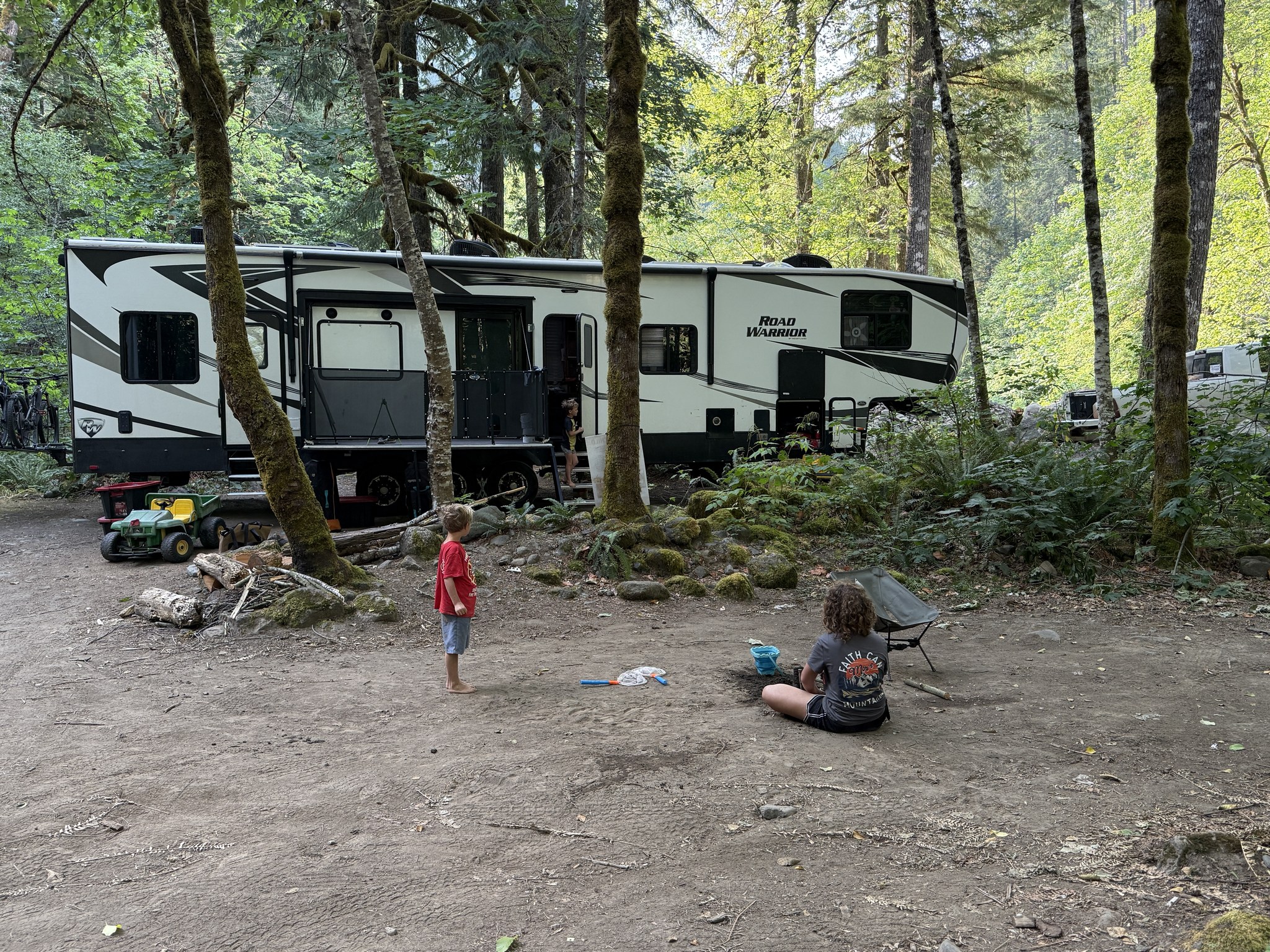 Children spend time outside their family RV in a shaded mountain campsite.