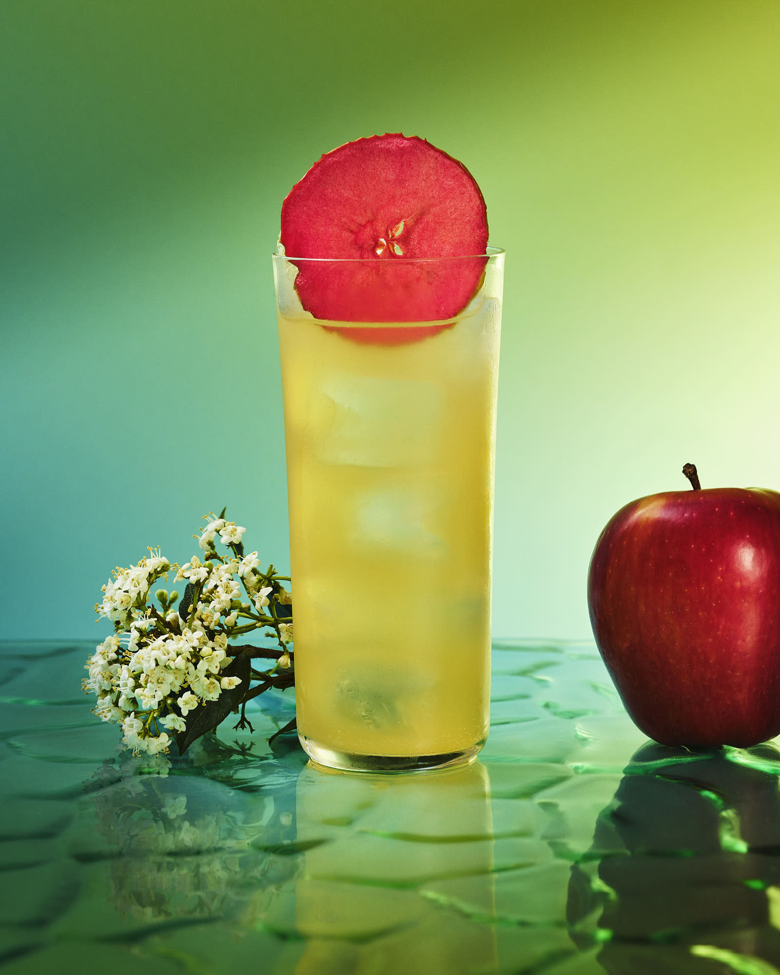 A refreshing drink in a clear glass, with a slice of fruit, accompanied by a red apple and green background.