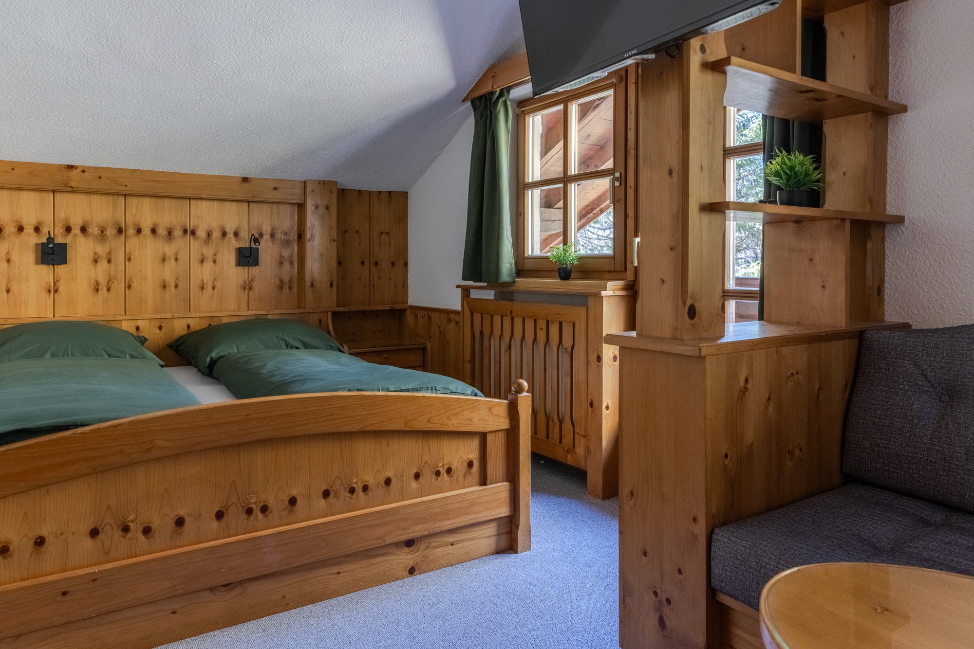 Ein gemütliches, rustikales Schlafzimmer mit Holzvertäfelung verfügt über ein bequemes Doppelbett mit grüner Bettwäsche, ein kleines Fenster mit grünen Vorhängen, das Tageslicht hereinlässt, und einen hölzernen Beistelltisch neben einem gepolsterten Sessel, was eine warme und einladende Atmosphäre schafft.
