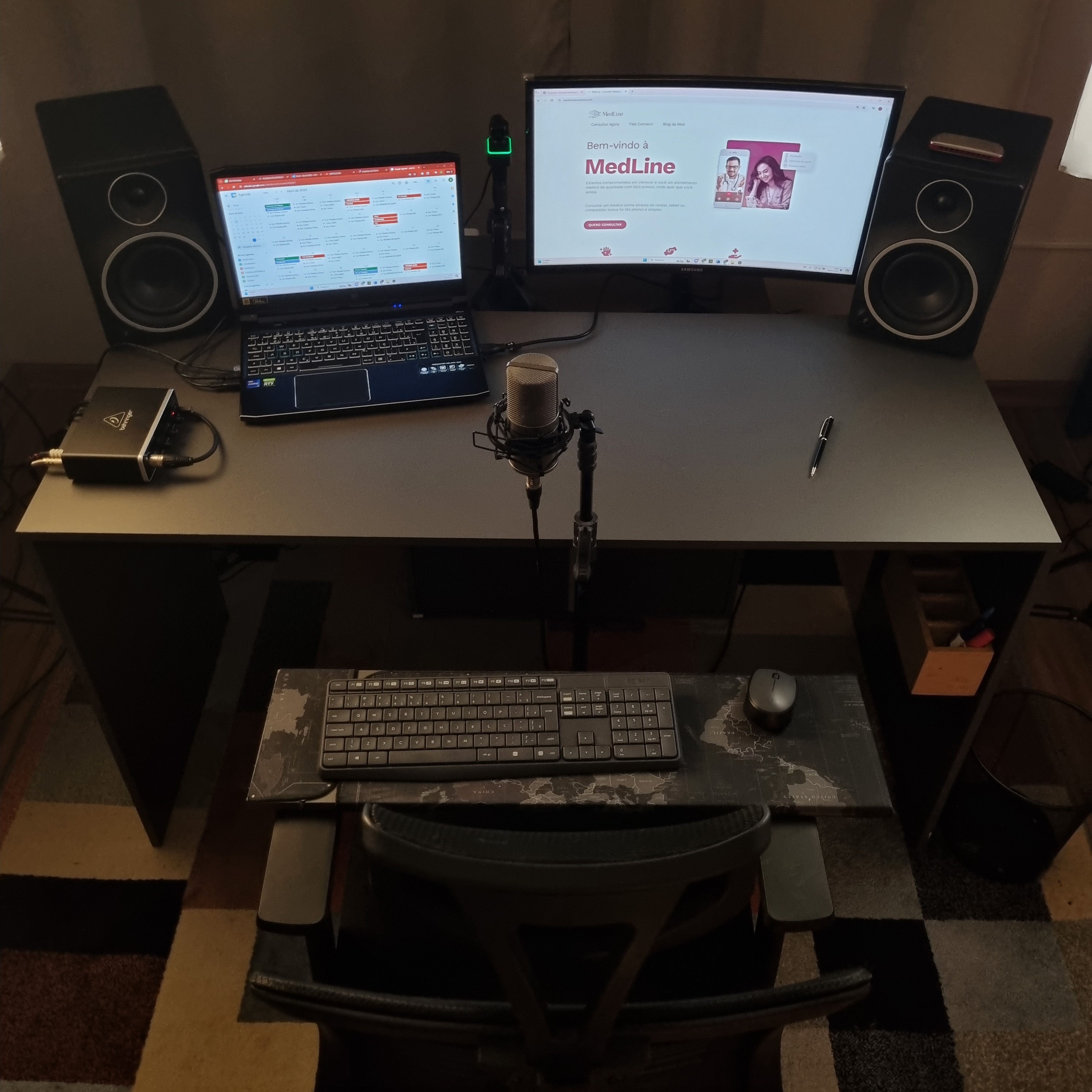 Microfone condensador MXL 770 em um suporte antivibração (shock mount) sobre um pedestal, conectado por um cabo XLR. Ao fundo desfocado, um monitor de computador exibe o texto "MedLine" e a imagem de uma pessoa, com cortinas visíveis na parte superior.