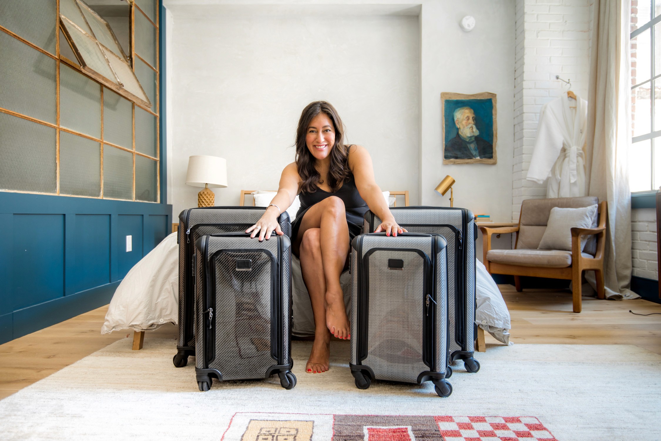 A woman in a black dress sits on two large suitcases in a bright room with white walls and blue wainscoting.