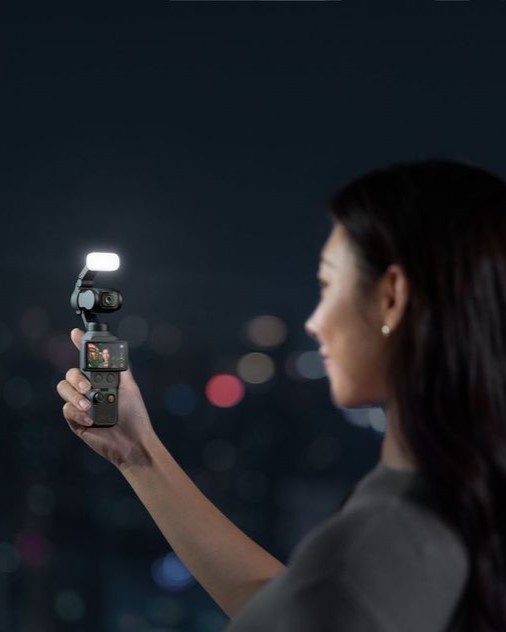 A woman holding a handheld camera with a light on top, standing against a night city skyline.