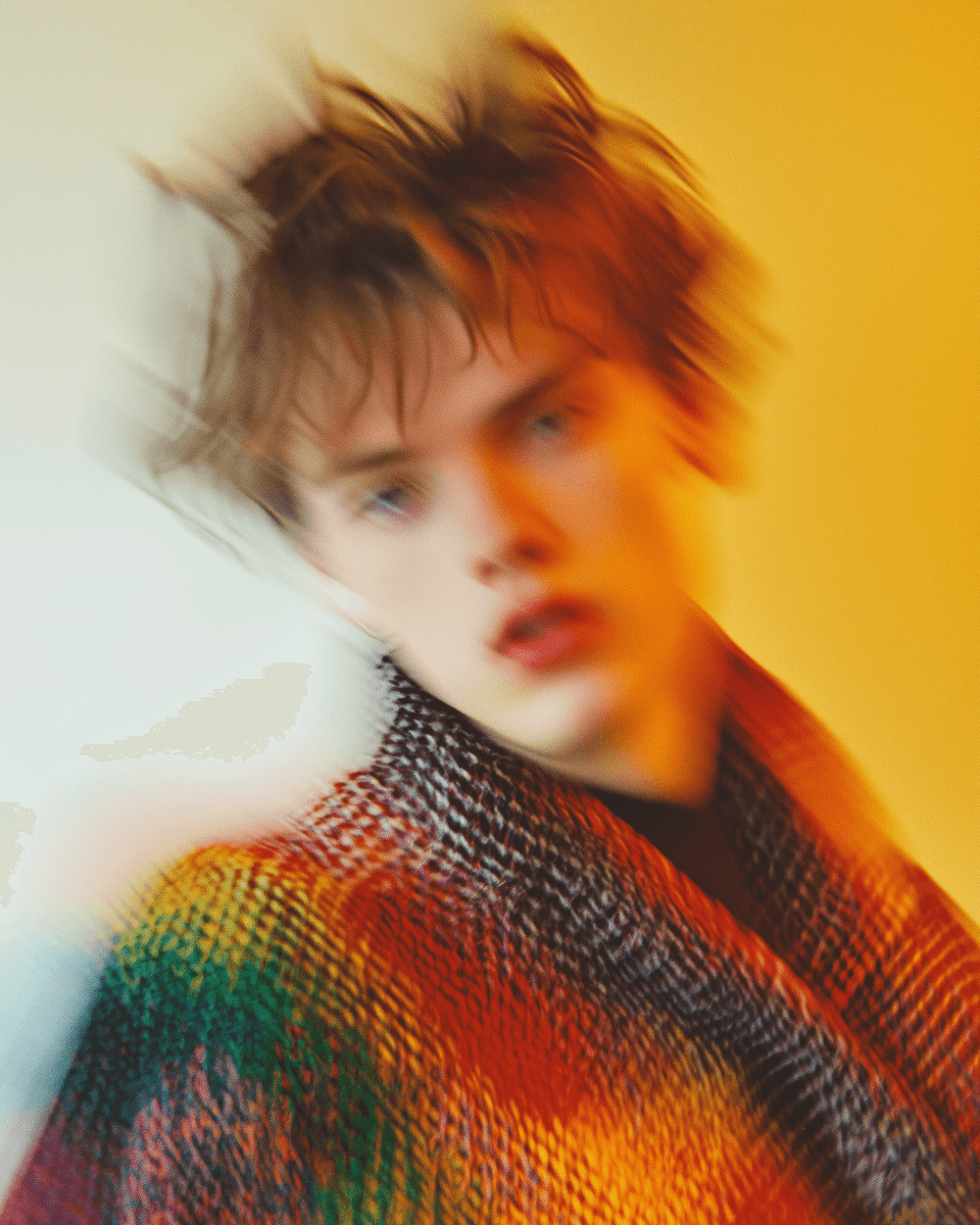 A blurred portrait of a person with messy hair, wearing a colorful sweater against a bright, warm background.