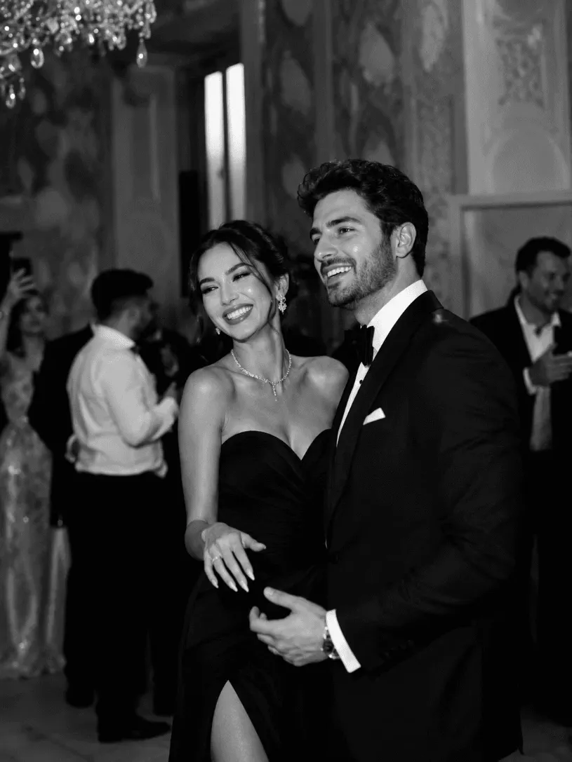 A joyous couple dressed in elegant formal attire shares a moment on the dance floor during a sophisticated evening event, with a chandelier and well-dressed guests in the background.