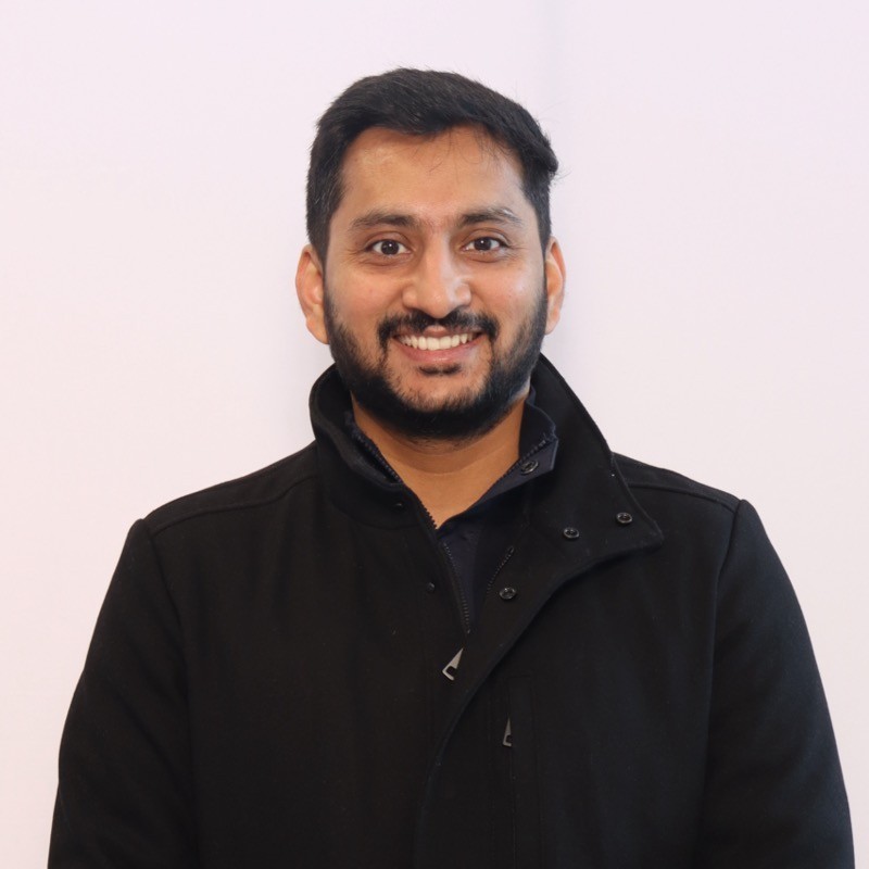 A professional chef, wearing a white chef's coat and toque, stands with his arms crossed, smiling warmly at the camera, with warm pendant lights and a kitchen area blurred in the background.