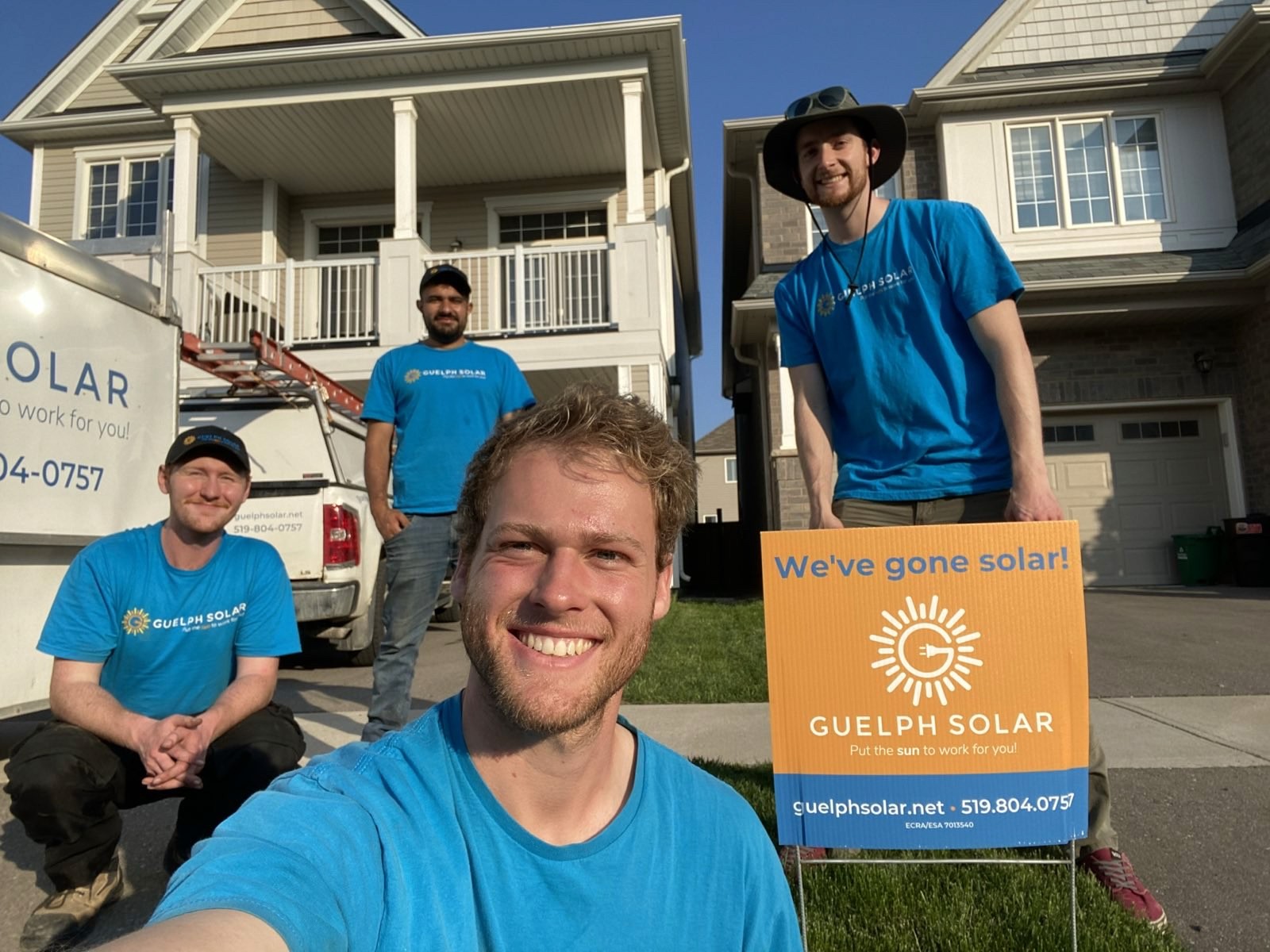 Four smiling guelph solar installers in blue shirts pose outdoors in front of a house, with a delivery box nearby.
