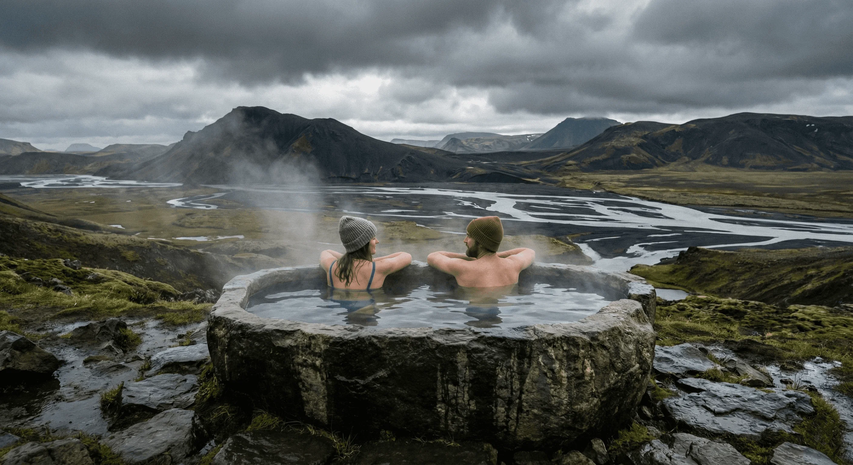 Two people relaxing in a circular natural hot pot surrounded by a rugged mossy landscape.