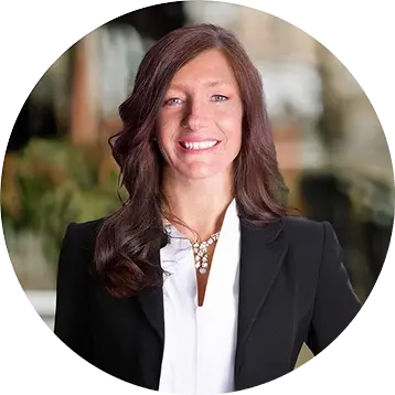 A professional woman with long brown hair smiles, dressed in a black blazer over a white shirt, against a blurred background.