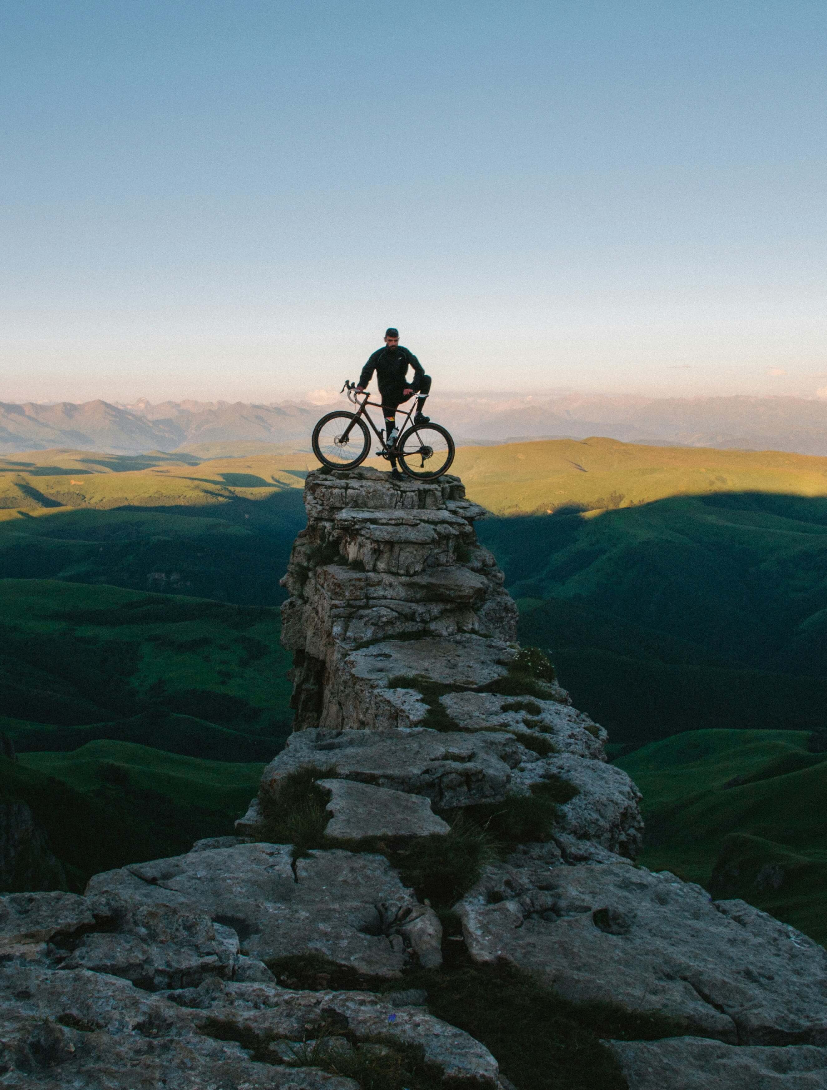 Cycliste avec son vélo au sommet d'une falaise vertigineuse, contemplant un panorama de montagne spectaculaire.