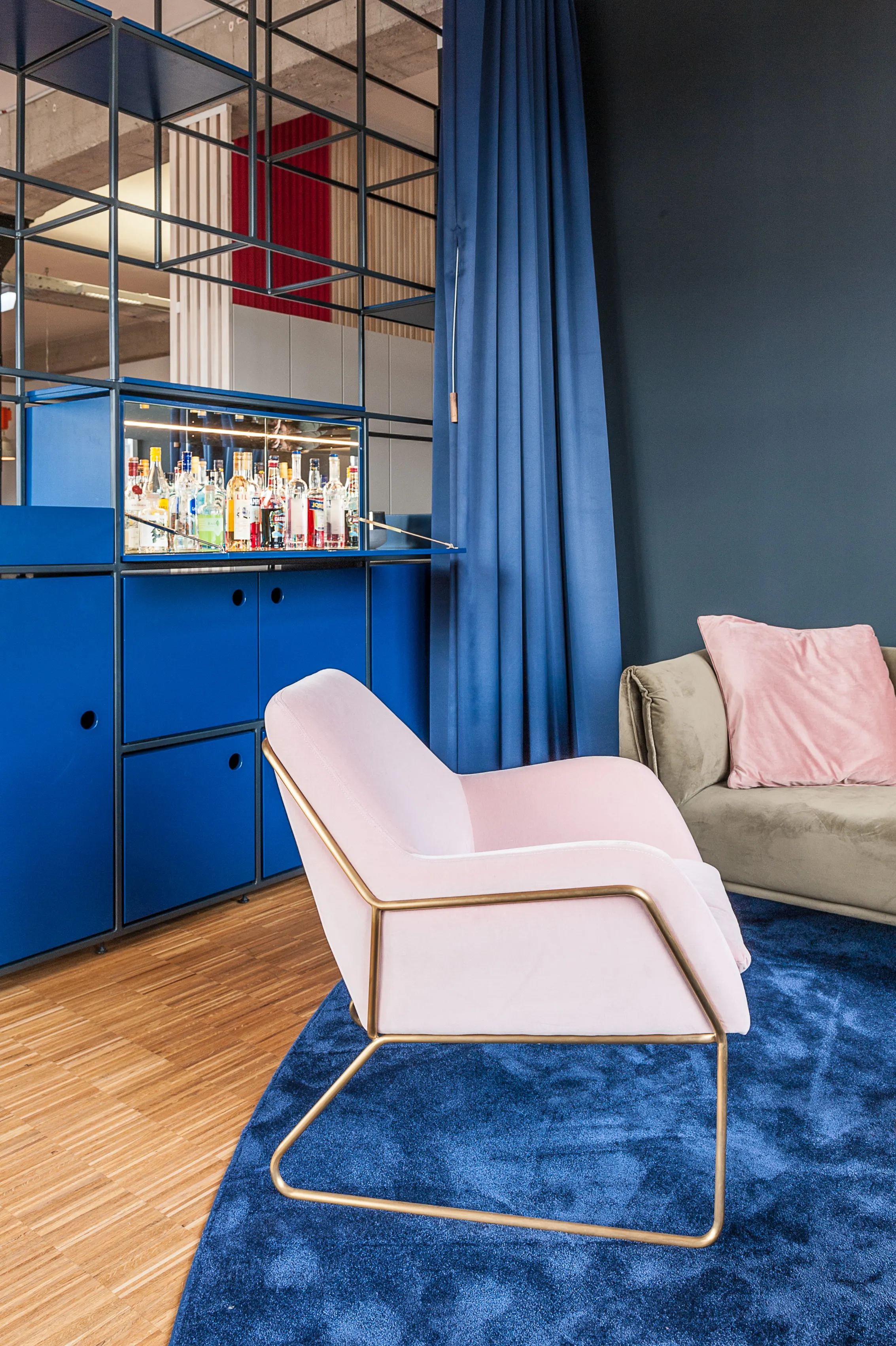 Pale pink armchair with brass frame on a deep blue carpet at BASILICOM, next to a beige sofa with pink cushion and a blue bar cabinet stocked with bottles behind a navy curtain.