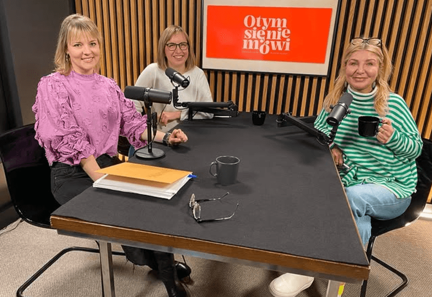 Three women sitting at a table with microphones, smiling and engaging in a podcast recording session.