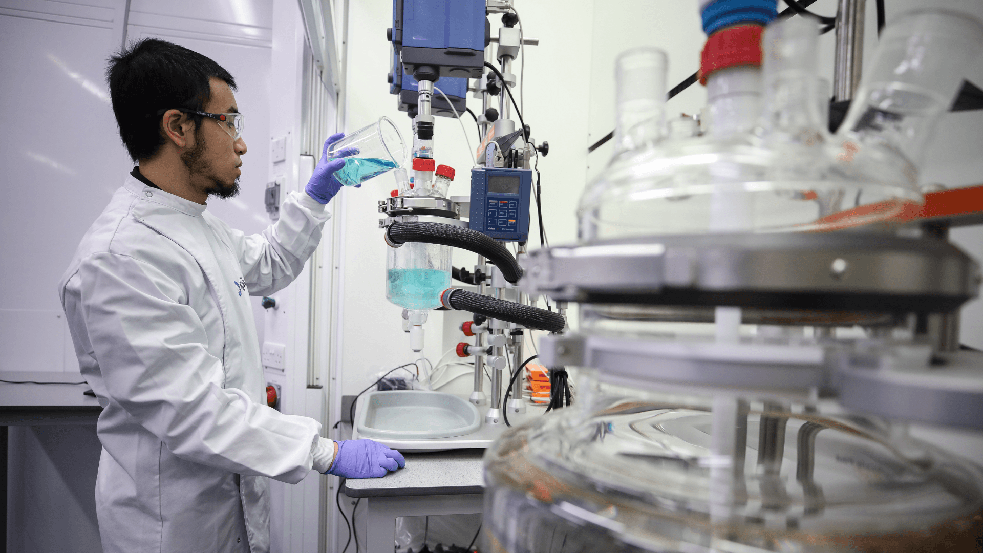 CPI scientist adding light blue liquid into into a glass reactor vessel.