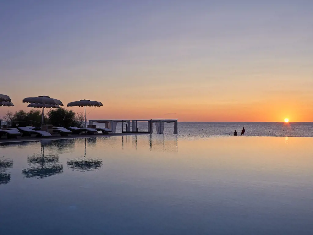 Sonnenuntergang über dem Meer mit Schwimmbad, Sonnenschirmen und Liegen, zwei Personen im Wasser.