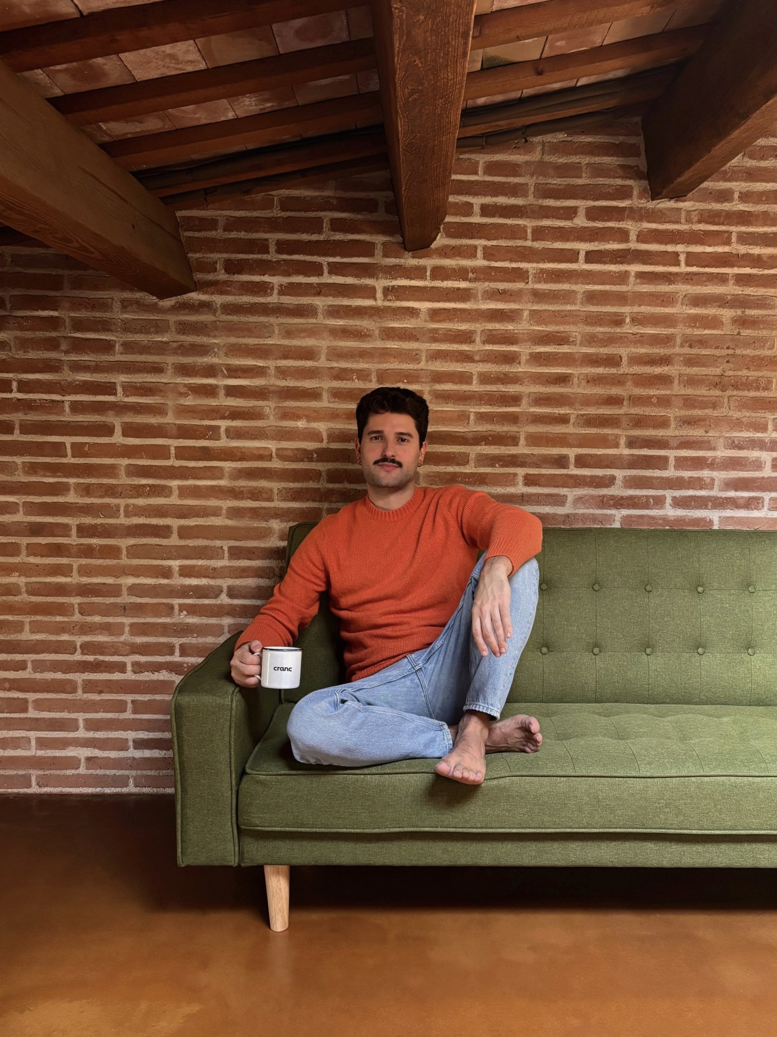 A person sits cross-legged on a green couch, holding a mug, in a cozy room with a wooden ceiling.