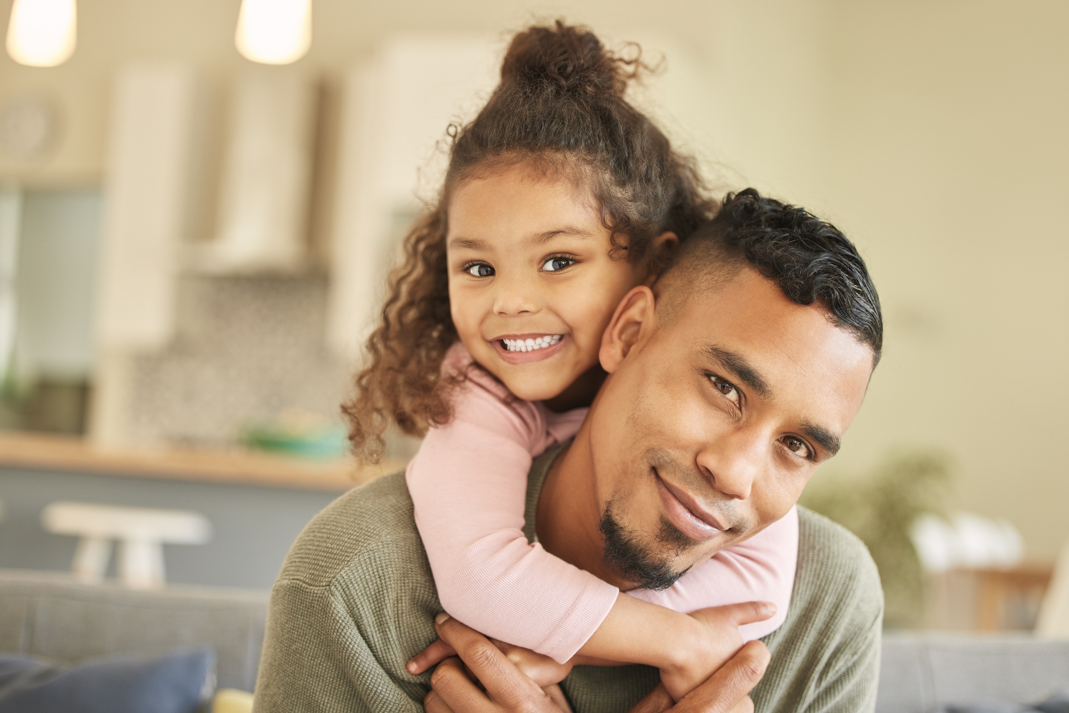 Imagem de um pai jovem e sua filha em um momento afetuoso em casa. A menina, com um sorriso alegre e cabelos cacheados, está abraçada ao pescoço do pai, transmitindo carinho e cumplicidade. O pai sorri levemente, demonstrando felicidade e amor. A cena é iluminada e acolhedora, retratando o vínculo familiar e a alegria da convivência entre pai e filha em um ambiente doméstico.