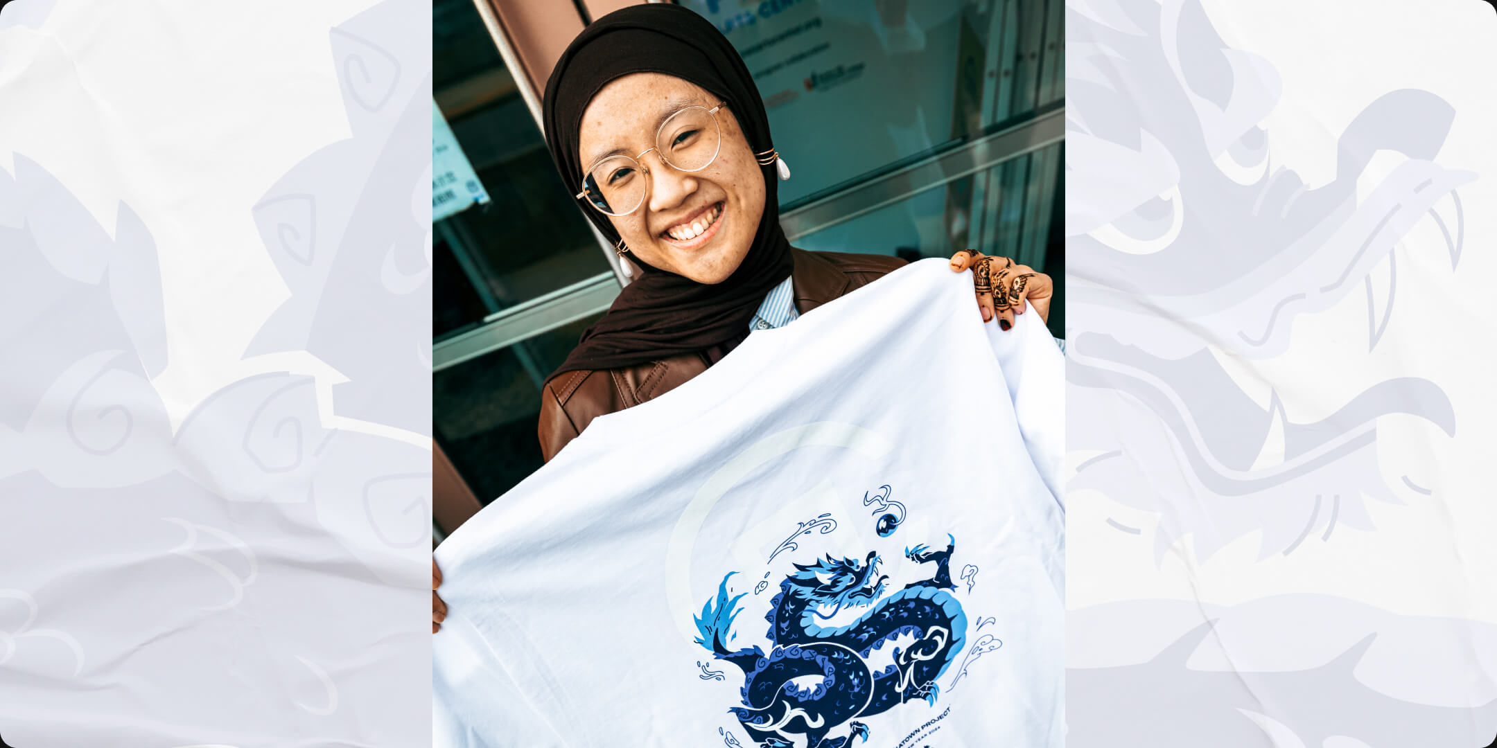 Artist Victoria Lai is smiling while holding up a white T-shirt featuring a detailed blue dragon design. She is wearing glasses, a brown jacket, and a black hijab. The background includes partial views of buildings and a subtle dragon graphic overlay.