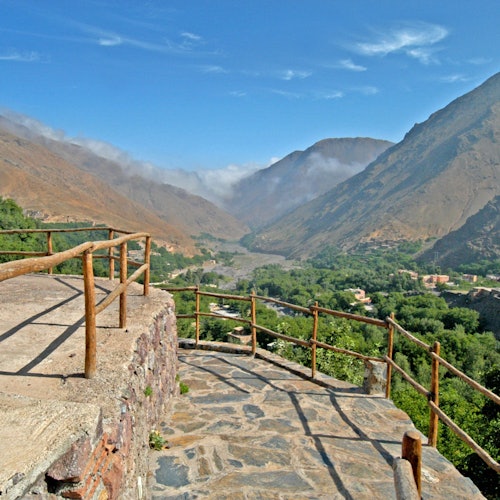Stone terrace with wooden railings overlooking lush, green valley between mountains and under a blue sky with scattered clouds.