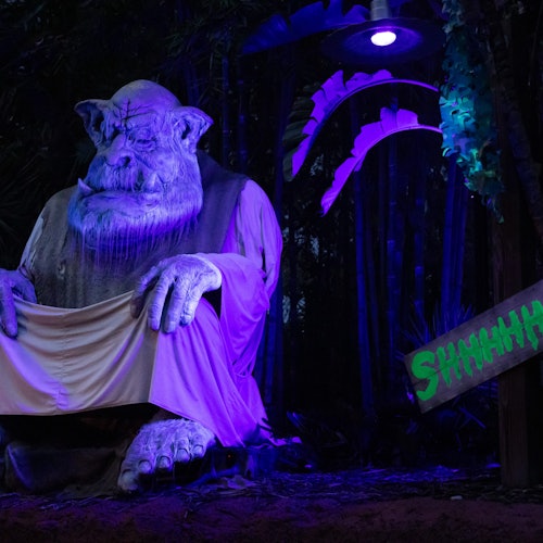 A large statue of a sitting troll under purple lighting with a "Shhhhhh" sign nearby in a forested area.