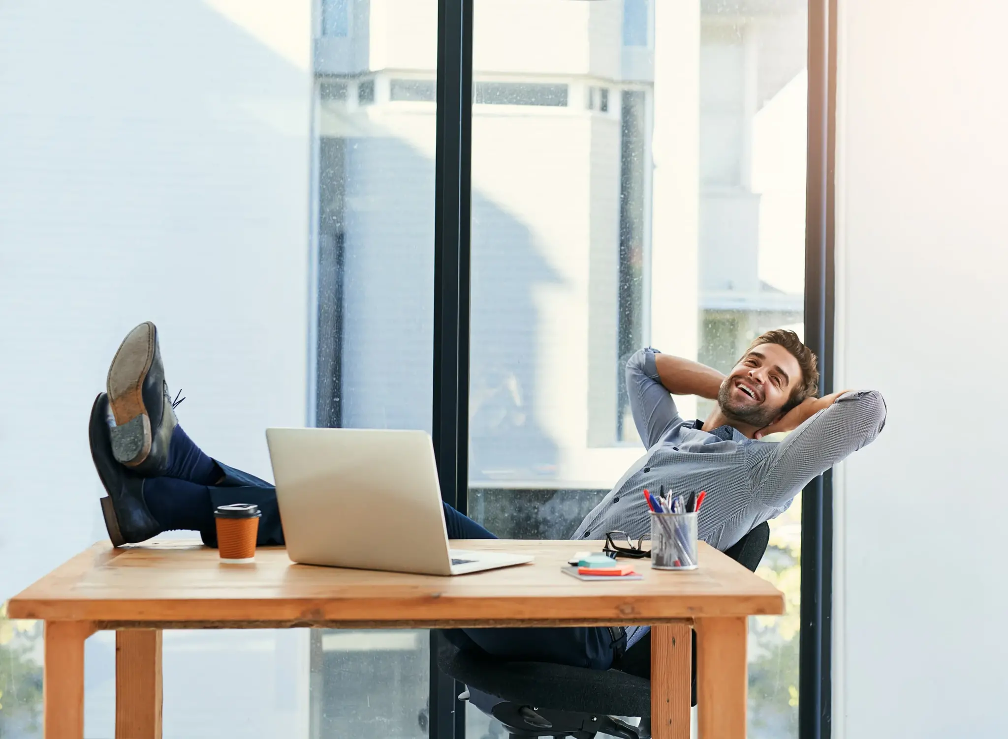 A relaxed businessman leans back at his desk with his feet up, smiling confidently beside a laptop and coffee cup in a bright modern office. The image represents stress-free investment financing through Debt Service Coverage Ratio (DSCR) loans from Chris Lewis Home Loans.