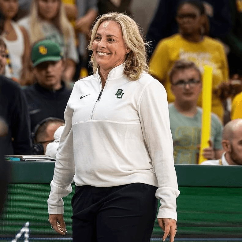 Nicki Collen in a white Baylor University pullover and black pants stands courtside at an indoor sports event. Spectators in yellow and green shirts are visible in the background.