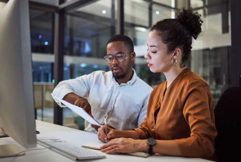 Two professionals collaborating at a computer screen, one pointing at data and the other taking notes, representing agency audit and compliance review