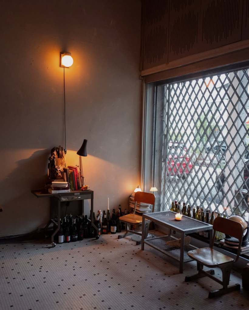 Cozy interior of a small cafe featuring a table and two chairs, a dimly lit lamp, and a collection of wine bottles along the window sill.