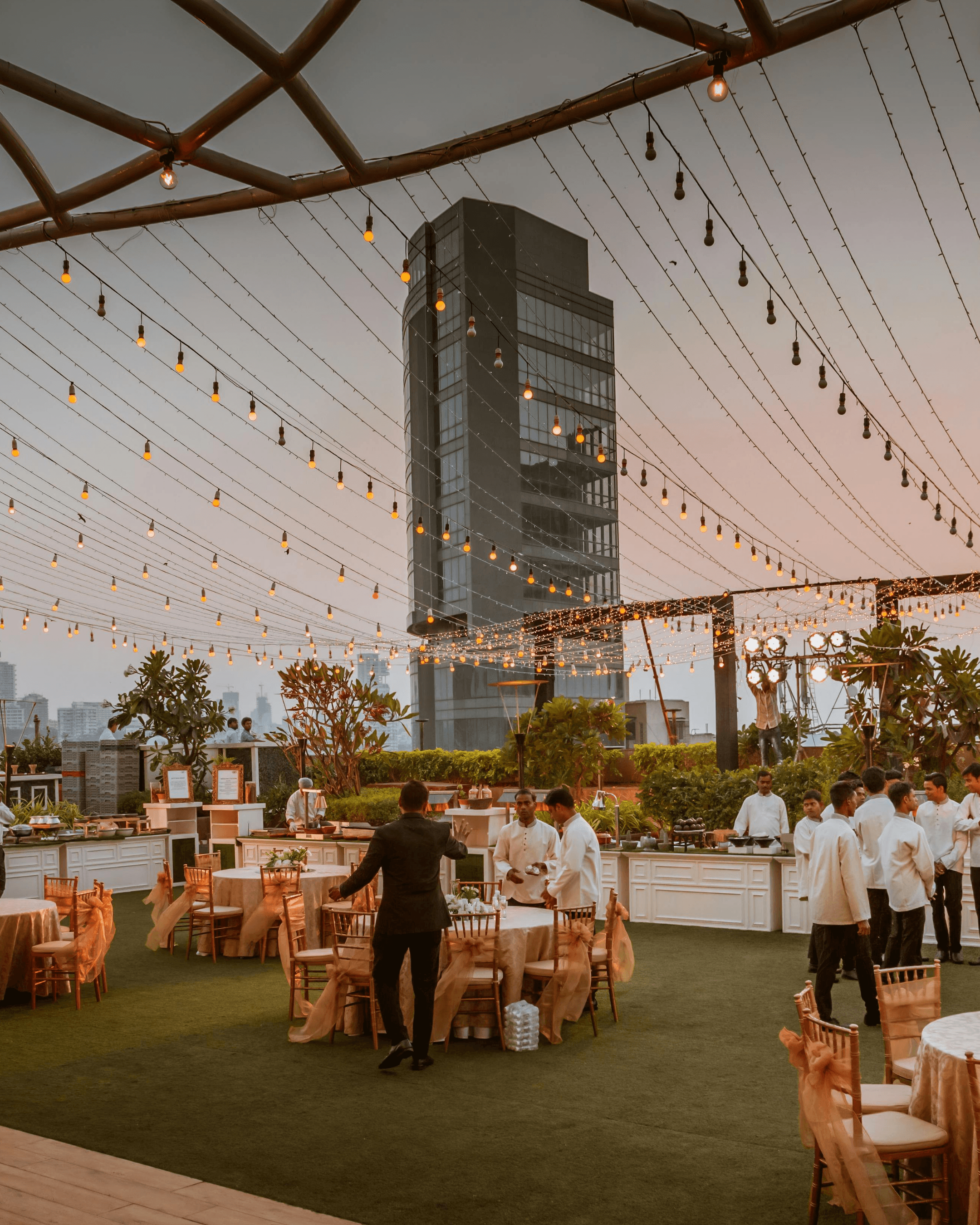 Evento al aire libre al atardecer, con mesas, buffet e iluminación colgante.
