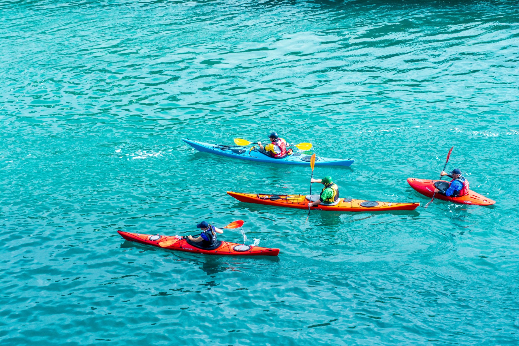 Four kayakers in vibrant red, orange, and blue kayaks paddle on clear turquoise water. The scene conveys a sense of adventure and leisure.
