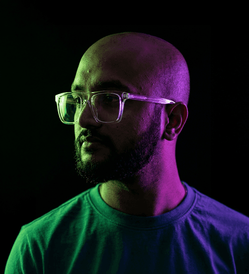 Videographer Vamshi L, bald with a beard and glasses, illuminated by teal and purple lighting.
