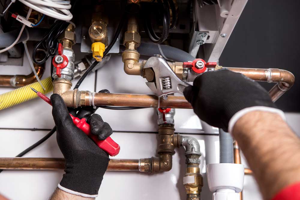 A plumber wearing black gloves uses a wrench to adjust copper pipes near a heating system, part of a heating installation in Asheville. Various tools and connectors can be seen, along with a yellow flexible gas pipe and red-handled valves. The plumber holds a red screwdriver in the other hand.