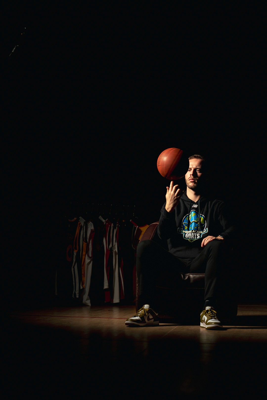 Athlète assis, manipulant un ballon de basket, dans une salle faiblement éclairée avec des maillots visibles.