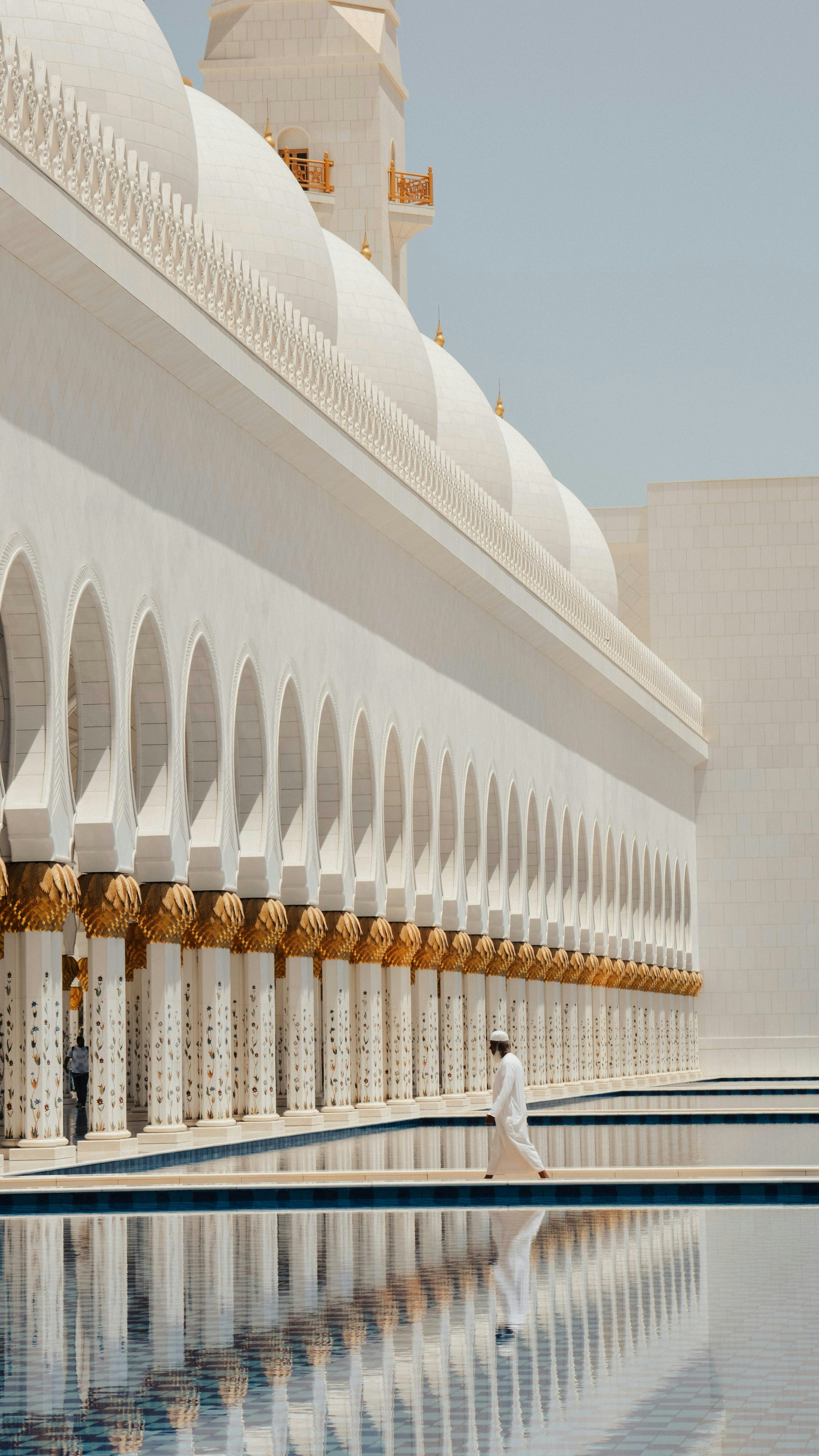 A person walks beside a white mosque reflecting in water.