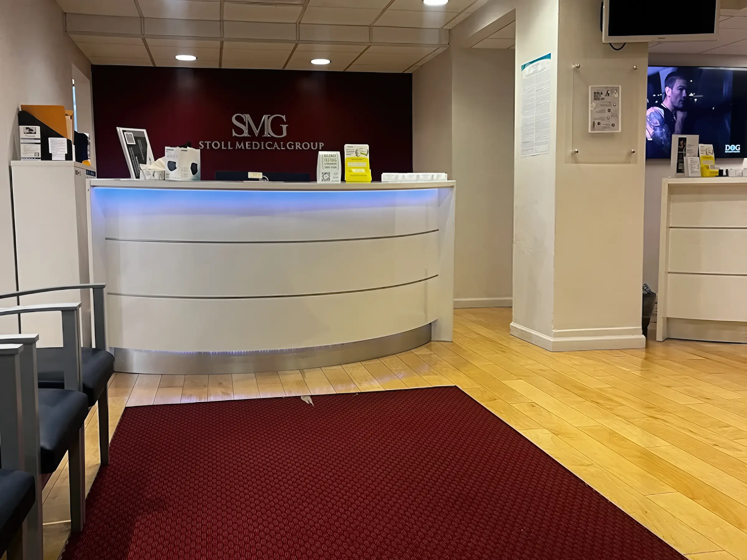 Hardwood flooring in a Stoll Medical Group front desk area with a curved illuminated reception counter, SMG branding wall, and waiting area seating.