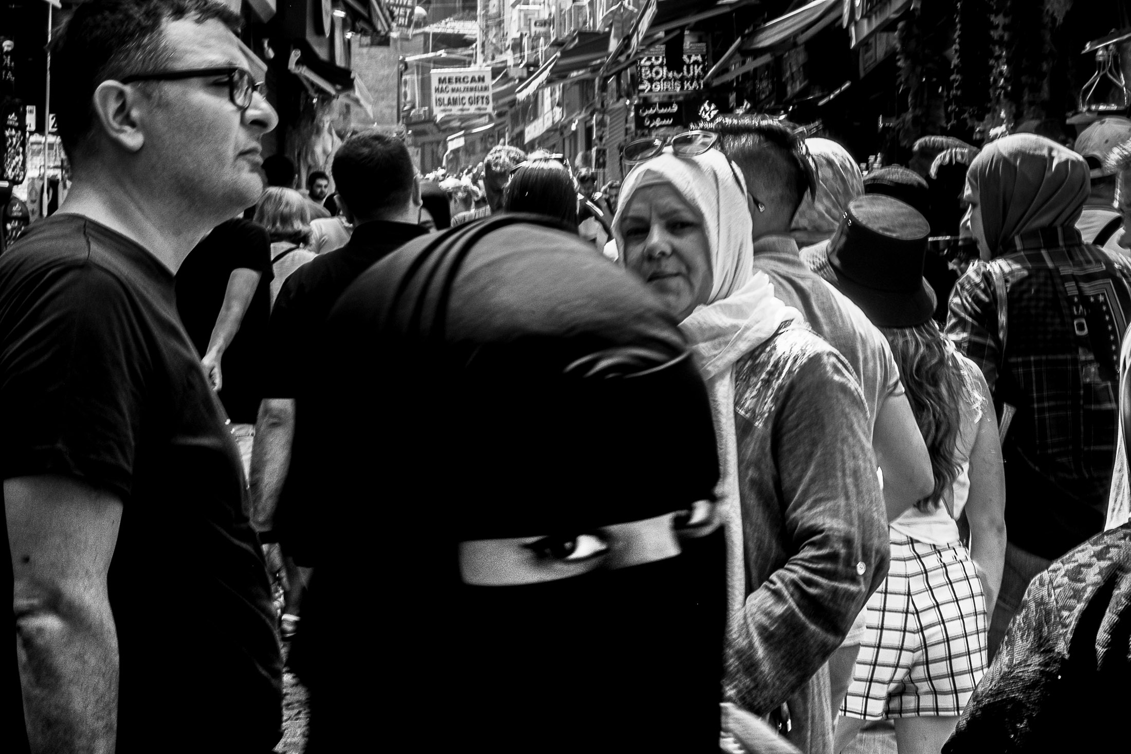 Crowded street market scene with layered faces and a woman wearing a headscarf looking toward the camera.