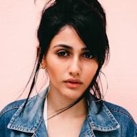 Portrait of a woman with dark hair styled up with loose strands, wearing a denim jacket, against a solid light pink background.