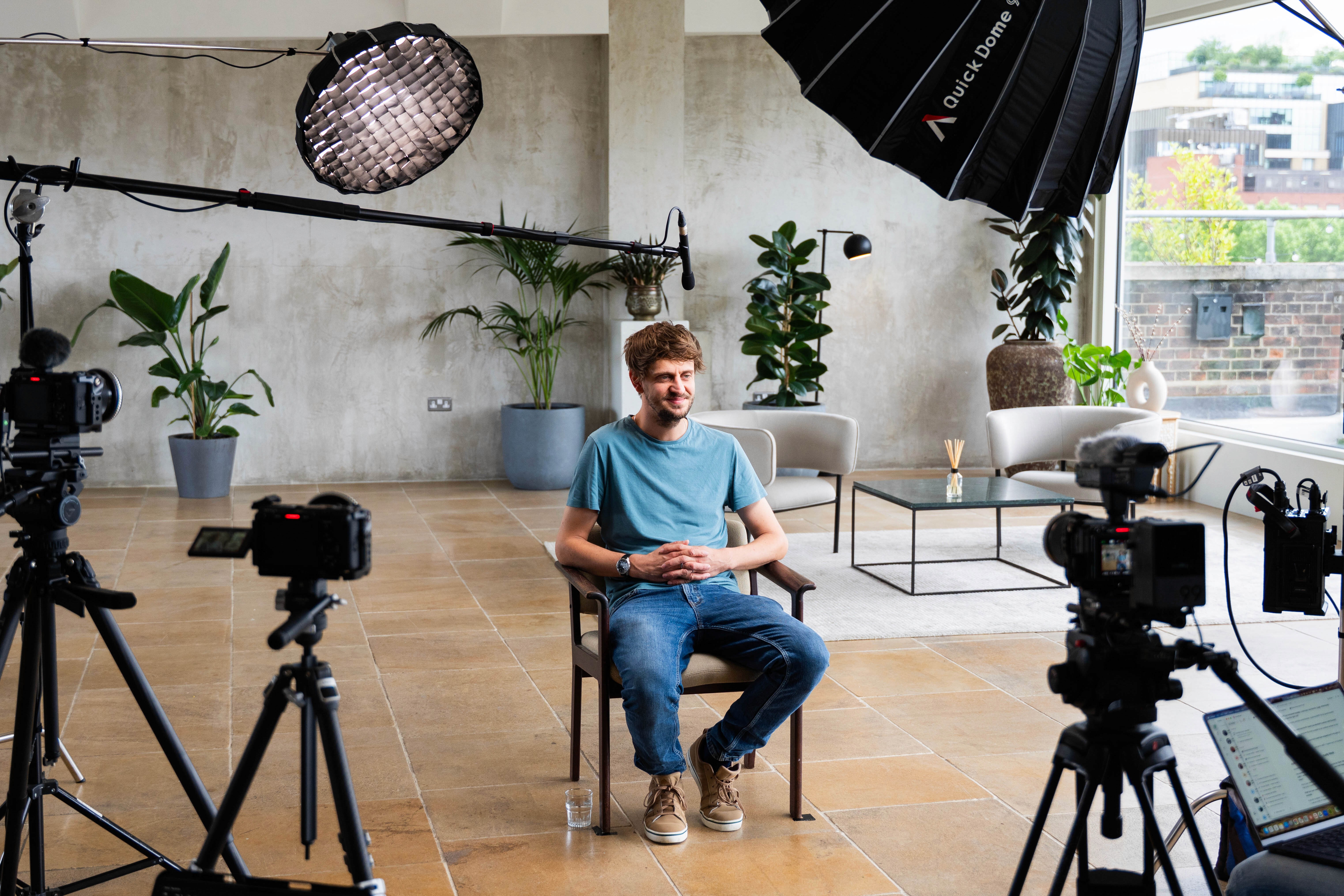 A man in a blue t-shirt and jeans seated in a wooden chair during a professional video shoot, surrounded by two cameras on tripods, softbox lighting rigs, and a boom microphone, in a bright studio space with potted plants and floor-to-ceiling windows.