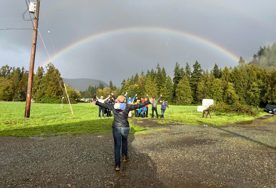 Community members raising their arms beneath a rainbow over the fields at Rooted Northwest.