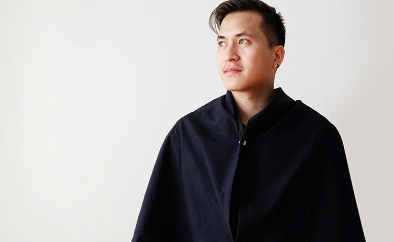 A person with short dark hair and light skin stands against a plain off-white background. They are wearing a dark navy blue poncho with a zipper closure at the neck. The poncho covers their shoulders and upper body. Their gaze is directed to their left, and their expression is neutral.